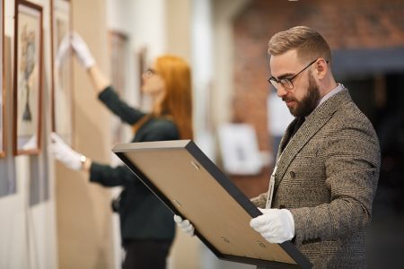 Art manager in suit and in gloves standing an looking at picture in his hands while woman hanging painting on the wall at gallery